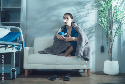 Canvas Print Patient Receiving Inhaler Treatment: A young woman sits on a couch wrapped in a blanket, using an inhaler in a room that appears to be a hospital or clinic