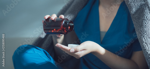 Foto Sick Woman Taking Medicine: A woman wrapped in a blanket, sitting on a bed, pours pills from a bottle into her hand