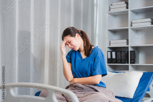 Fotografía A Patient in Distress: A young Asian woman in blue scrubs sits on a hospital bed, looking unwell and holding her forehead in discomfort, conveying a sense of pain and worry