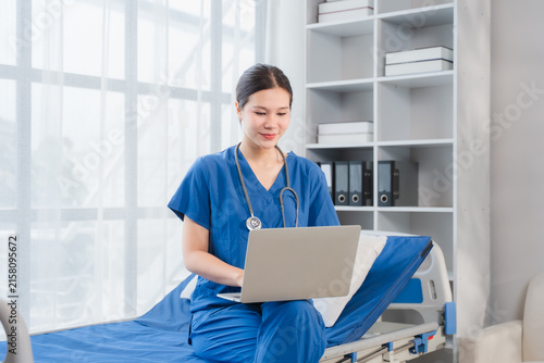 Canvastavla Nurse in Clinic: A focused nurse in blue scrubs sits on a patient bed, attentively working on a laptop with a stethoscope around her neck, in a bright, modern clinic setting