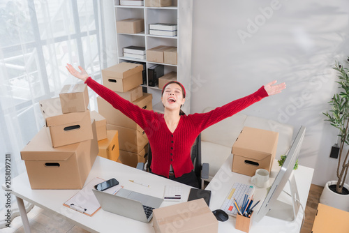 Photography Startup Success: An overjoyed woman sits at her desk, surrounded by cardboard boxes, arms outstretched in a moment of pure elation, celebrating the success of her burgeoning online business
