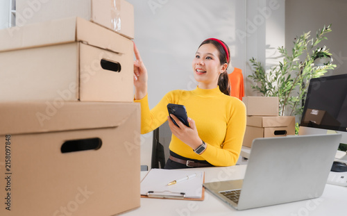 Quadro su tela Online Retailer Triumph: A smiling woman, an online shop owner, organizes inventory, surrounded by cardboard boxes, phone in hand, and laptop on desk, poised for success
