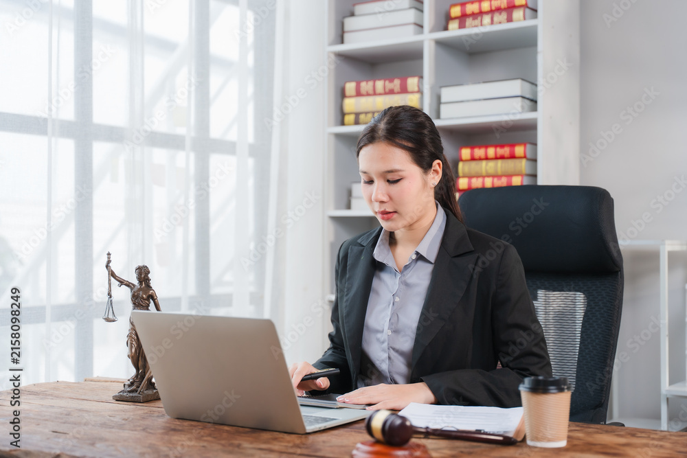 Fototapeta premium Lawyer Working in Office: A focused female lawyer in a professional suit works diligently on her laptop at a wooden desk, surrounded by legal books and a statue of justice.