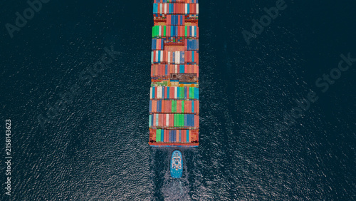 Aerial view of a large container ship loaded with colorful cargo containers being guided by a tugboat through the deep blue waters of Piraeus Harbour in Athens, Greece.