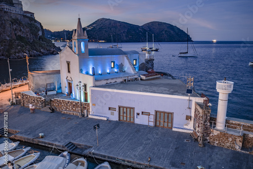 Aerial view of the illuminated Church of the Souls in Purgatory situated by the harbor with moored boats and calm sea at dusk in Lipari, Sicily, Italy.