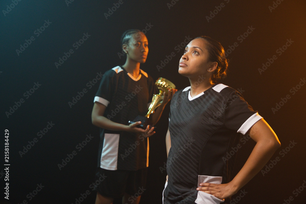 Fototapeta premium African American female teammates posing in studio wearing black-white jerseys and gold boot trophy