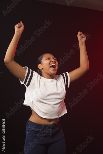African American woman raising arms, shouting in studio wearing white cropped tee lit by red rim