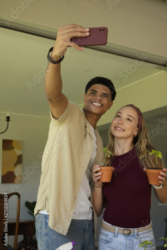 Diverse couple taking selfie in home dining area holding terracotta pots and maroon smartphone