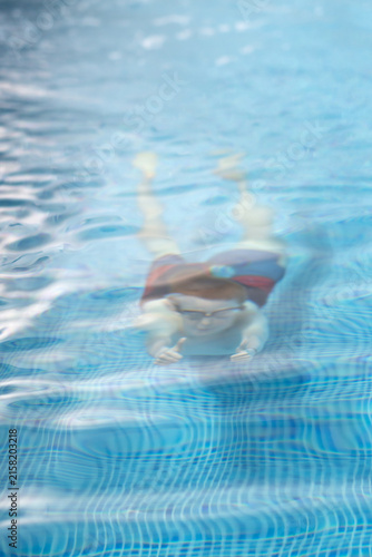 A young athlete in swimming goggles comes out of the water making hand signals while practicing diving in a clear blue pool.