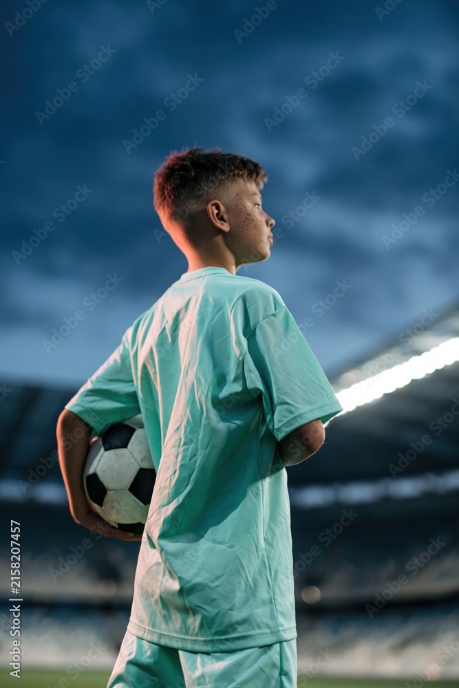 Naklejka premium Young soccer player in stadium at night holding a ball looking to the side for sports background poster and banner design for youth training