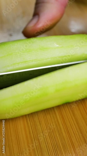 A close-up shot captures the precise movement of a chef's hands using a sharp knife to slice a peeled green cucumber into thin pieces on a wooden cutting board for a salad.