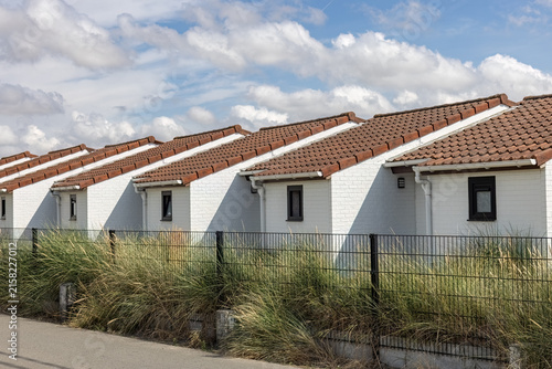 Row of identical white bungalows with red tiled roofs behind a metal fence and tall dune grass in Westende - Belgium