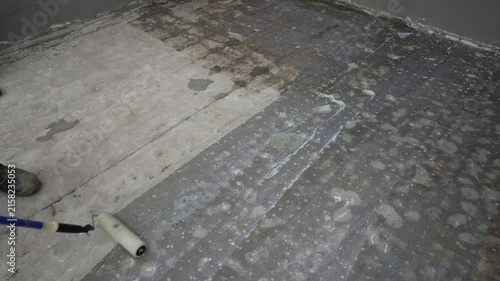 A worker uses a long handled roller to apply white primer onto a rough concrete floor. The action progresses from left to right showing coverage over stains and cracks in an indoor space