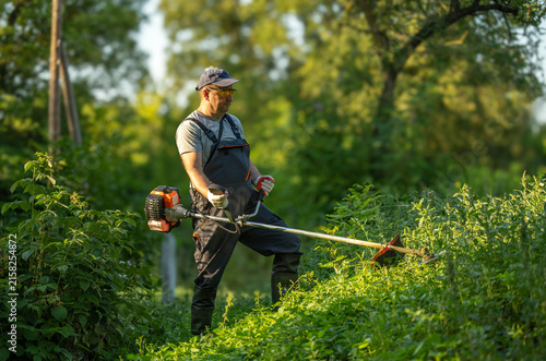 A gardener stands on a slope, operating a string trimmer to cut grass and weeds. The sun shines through trees, making the scene bright and active in the afternoon