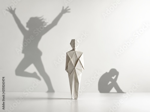 A still origami figure stands between two contrasting shadows, one joyful and energetic, the other withdrawn and sorrowful. The minimalist conceptual image symbolizes bipolar depression, emotional ext