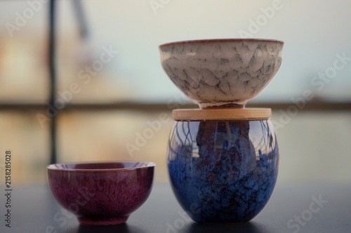 Three handcrafted ceramic vessels in burgundy, blue, and beige are arranged with one bowl balanced above a blue jar on a dark tabletop. The image represents equilibrium, playful creativity, and the