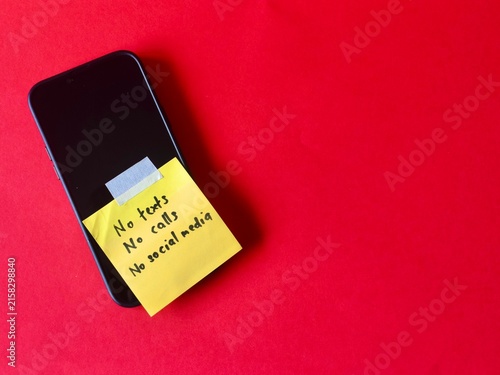 Fotografía A black smartphone positioned on a vivid red background with a yellow sticky note taped over the screen listing rules for a digital detox