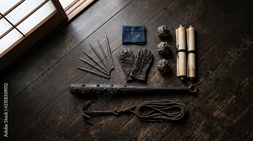 Authentic ninja gear displayed on dark wooden floorboards featuring sharp throwing knives leather gloves scrolls spiked club and metal grappling hook