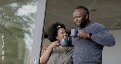 African American couple standing at sliding glass door sipping mugs and meeting gazes, connecting