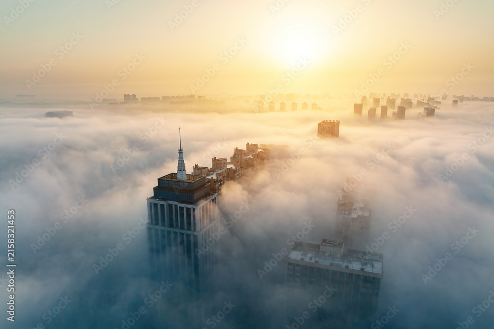 Fototapeta premium Tall urban buildings emerge from dense morning fog under the glow of a rising sun, creating a misty cityscape.