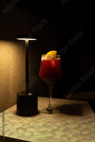 A vibrant red craft cocktail garnished with a citrus slice, illuminated by a modern warm table lamp in a dimly lit, upscale restaurant or speakeasy bar environment