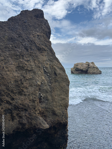 Large dark sea stack rock with marine life on a sandy beach looking out towards offshore rock formations and crashing ocean waves. Dramatic coastal scenery under a cloudy sky