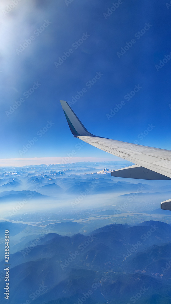 Fototapeta premium Airplane wing flying high above majestic snow-capped mountain ranges and layer of mist under a clear blue sky. View looking out of a passenger plane window during a sunny flight
