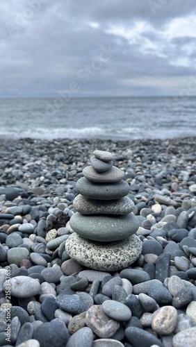 Balanced stack of smooth grey stones in a zen cairn on a rocky pebble beach. Serene coastal nature scene with gentle waves and an overcast sky background