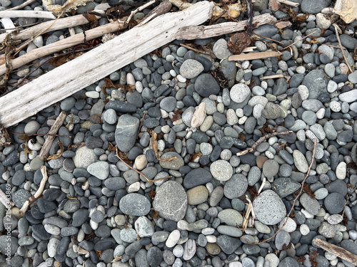 Top-down view of gray beach pebbles and weathered driftwood logs on a rocky shore. A natural textured background of stones, twigs, and dried seaweed washed up on the coastline.