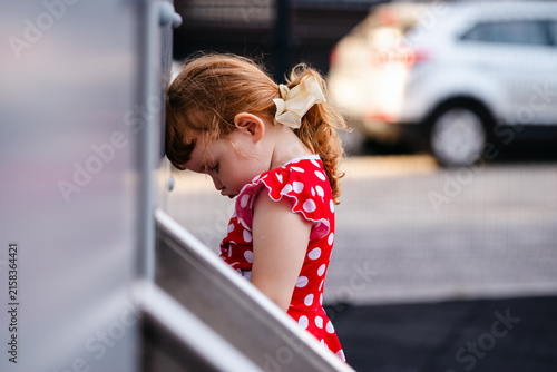 A sad, hurt red haired girl, standing alone by the street railing, expresses childhood suffering, loneliness, vulnerability, and the need for care in an urban environment.