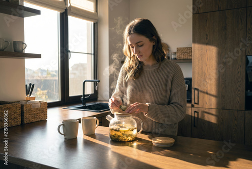 Young woman preparing herbal tea in a modern designer kitchen at sunset, surrounded by warm light and natural materials, creating a calm mindful wellness lifestyle scene