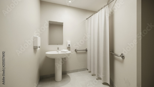 Accessible bathroom interior featuring pedestal sink and shower stall with safety grab bars in neutral tones with shower area enclosed white curtain