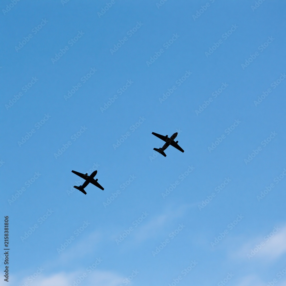 Fototapeta premium Two airplane silhouettes fly in formation against a clear blue sky with faint clouds below.