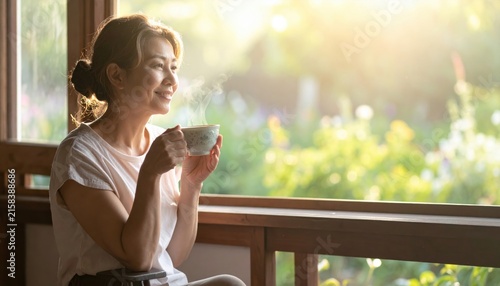 Smiling woman enjoying a warm cup of steaming coffee or tea by the window with sunlight and blurred greenery.