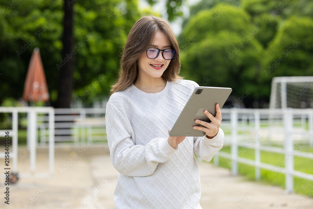 Fototapeta premium Young woman tablet outdoor park eyeglasses sweater smile digital technology leisure standing in green park while using tablet with relaxed expression near white fence and tree background