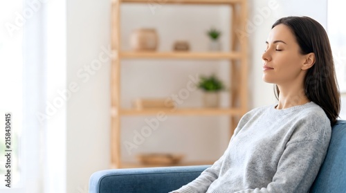 Young peaceful woman sitting on couch at home, eyes closed meditating and practicing deep breathing exercises for stress relief and mental health wellness