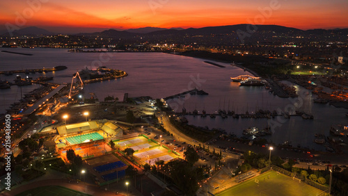 Aerial view of a marina with illuminated sports courts, including a swimming pool and tennis courts, situated along the coastline of Athens, Greece.