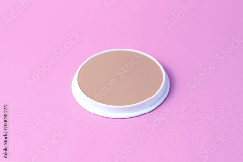 An open round cosmetic powder compact with cream-colored powder and a white case, displayed against a soft pink background.