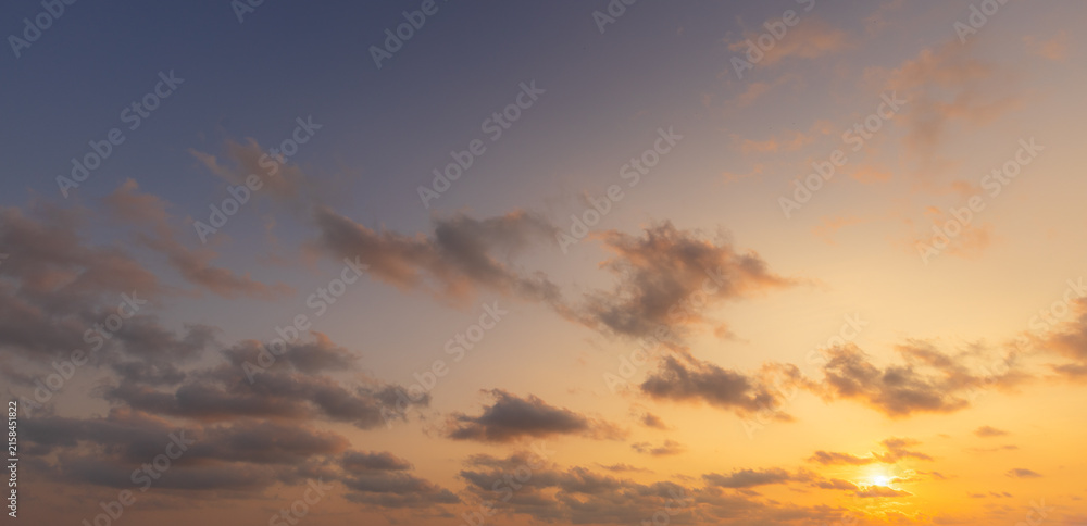 Fototapeta premium Gradient Sky Sunset,A beautiful, serene sunset sky transitions from a deep blue at the top to a warm, glowing golden-yellow near the horizon, filled with scattered, textured clouds. The sun is just.