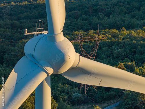 Aerial view of a wind turbine rotor and blades with transmission infrastructure in the background, illuminated by warm golden light in Marsanne, Auvergne-Rhone-Alpes, France.