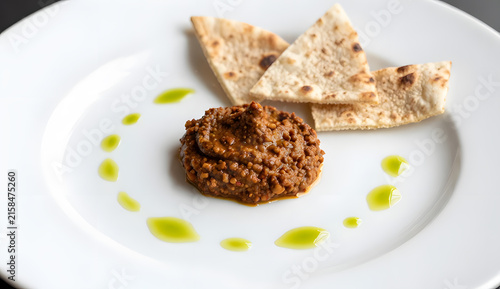 Harissa dip presented on a dramatically oversized white plate, tiny central mound of textured sauce surrounded by minimalist olive oil dots and three crisp flatbread shards, extreme close-up cropping 