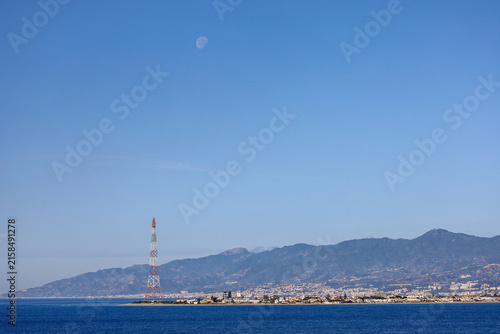 The historic red and white steel electricity Pylon of Messina at Torre Faro under a daytime moon, Sicily.