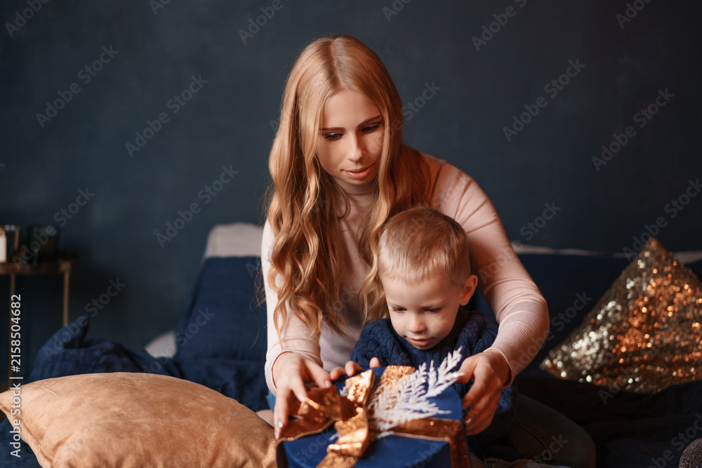 Naklejka premium Hands of a young caucasian mother and her little son unwrapping a blue present with a golden bow at home, festive surprise image for christmas gift campaigns.