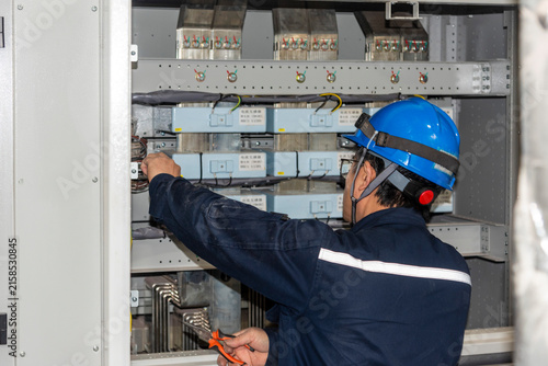 Shipyard electrician working on an open electrical control box inside the engine room of a newly built vessel, reaching toward electrical components during ship construction and commissioning.