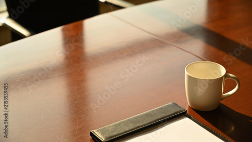 Glass cup of hot tea with infuser and yellow notepad on wooden conference table in sunny office