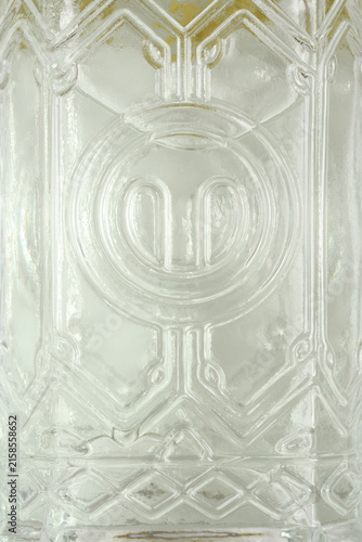 Full frame close up view of an embossed geometric pattern and decorative emblem on a vintage clear glass bottle.