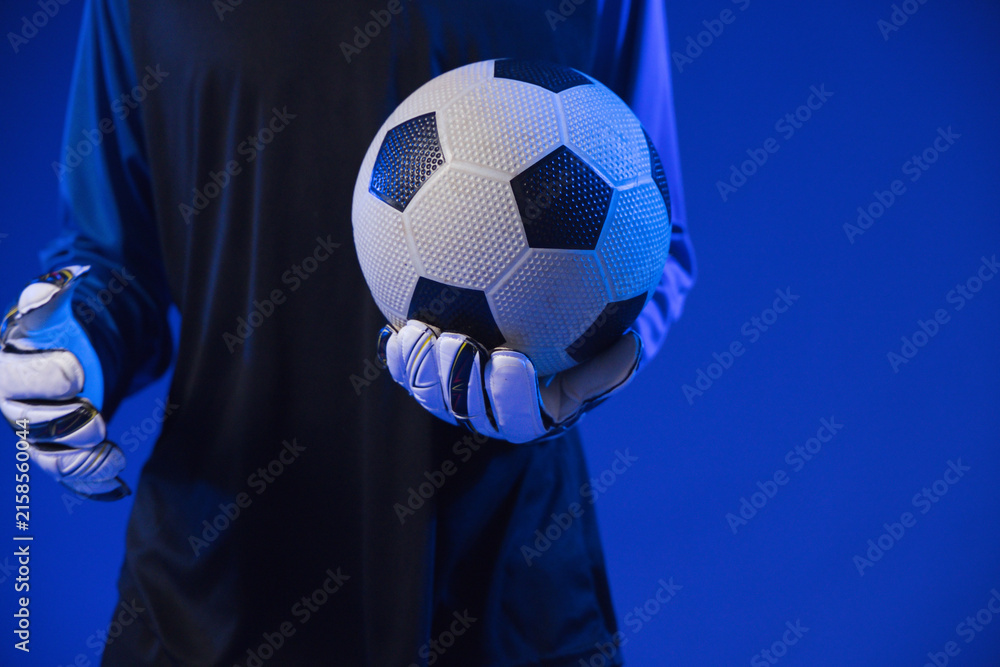 Fototapeta premium African American man holding black-white soccer ball at chest in blue studio, dark jersey, gloves