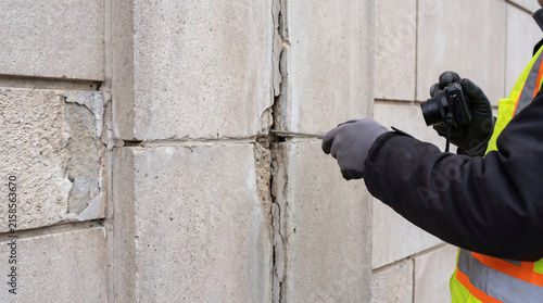 Building inspector examining structural crack and foundation damage in exterior concrete masonry wall with probe tool and camera
