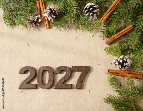 Year 2027 is framed by festive pine branches, frosted cones, and aromatic cinnamon sticks on a cozy linen background, symbolizing new beginnings and holiday cheer