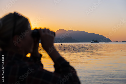 Defocused Woman standing on the waterfront in Marmaris, Turkey looks through binoculars toward the sea during golden hour at sunrise or sunset. Coastal promenade overlooks calm water and distant